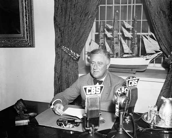 President Franklin Delano Roosevelt sitting at a desk with CBS and NBC microphones in the foreground.