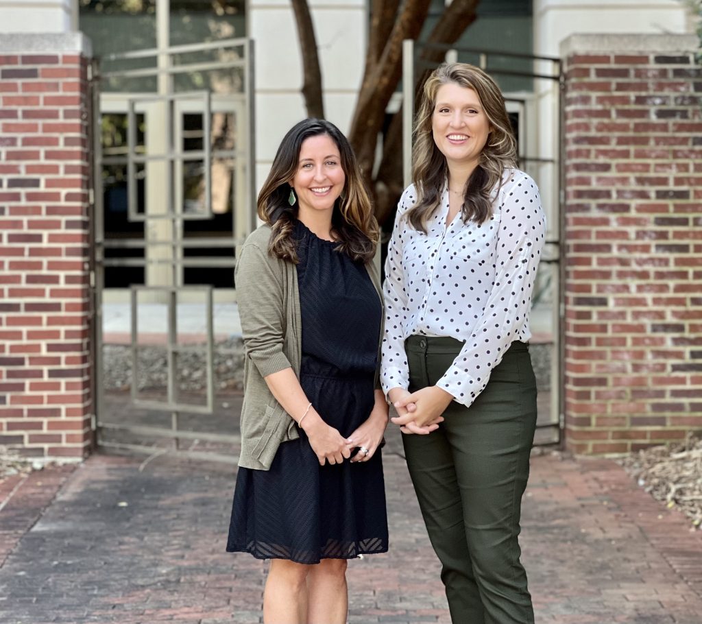 Lisa de Saxe Zerden, Ph.D., and Brianna Lombardi, Ph.D., stand in front of a gate near the Tate-Turner-Kuralt Building. 