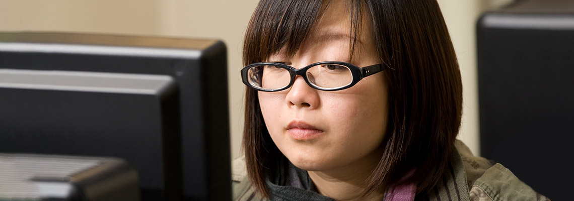 A young woman with glasses sits in front of a computer.