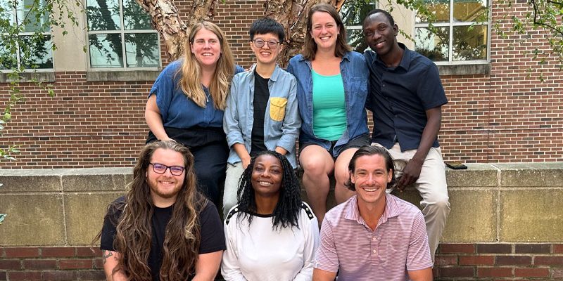 Seven students from the new 2023-2024 Ph.D. cohort pose for a photo from a bench and stone wall in front of the Tate-Turner-Kuralt Building on UNC-Chapel Hill's campus.