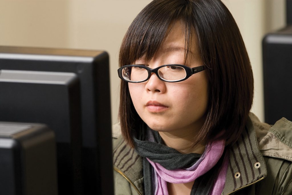 A young woman with glasses sits in front of a computer.